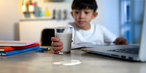 Image: A student holding a glass of milk with a spill near a school laptop — highlighting the need for device protection.